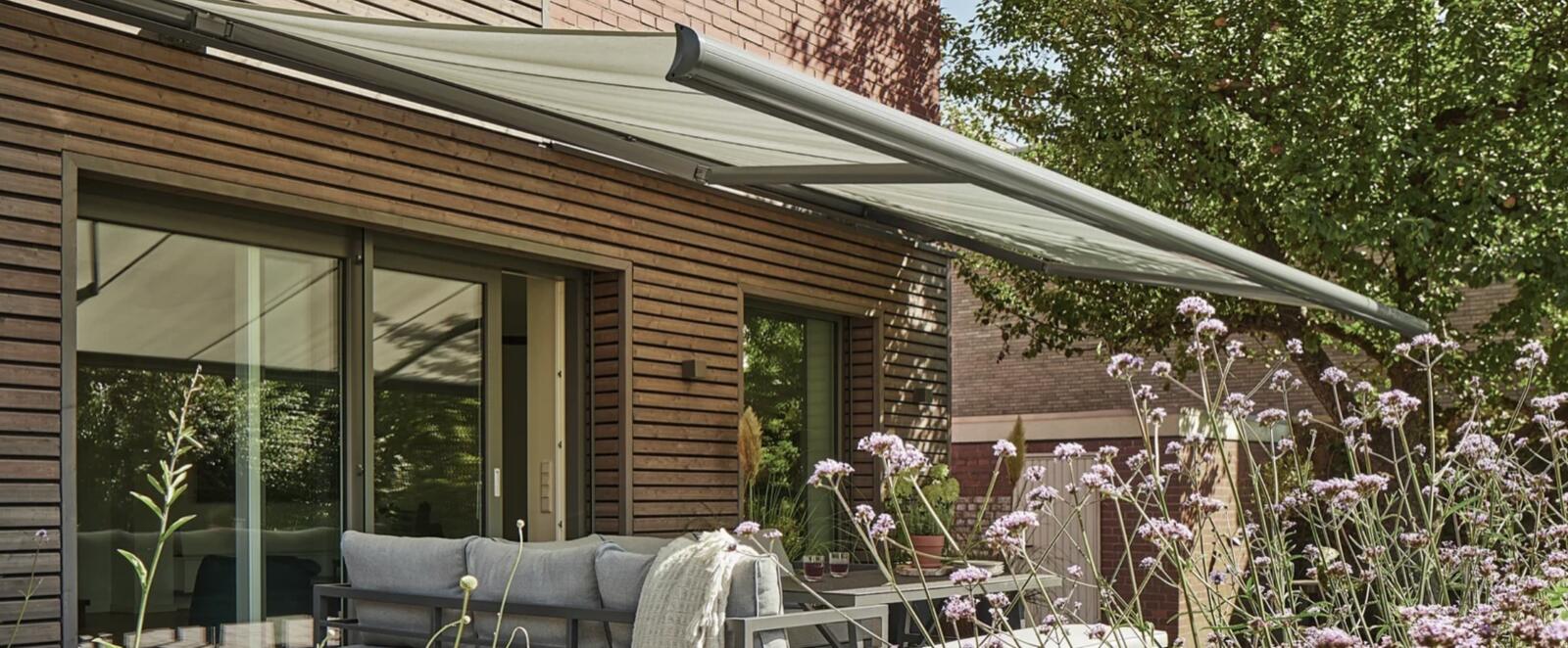 Ausgefahrene Kassettenmarkise in Olivgrau über einer Holzterrassenfassade mit Lounge-Möbeln und blühendem Garten