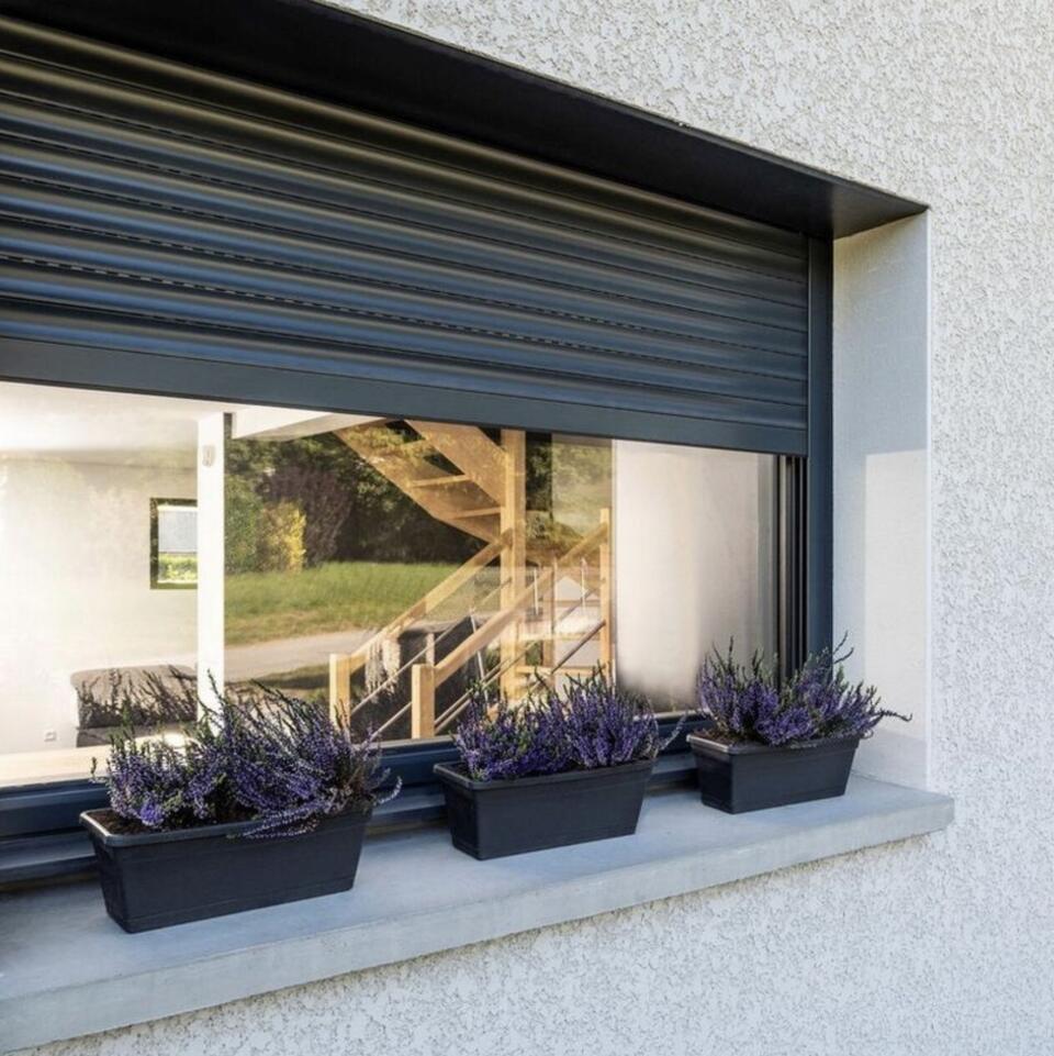 Anthrazitfarbener Rollladen mit halb geöffneter Lamelle an einem Wohnhausfenster mit Lavendelblumen auf dem Fensterbrett